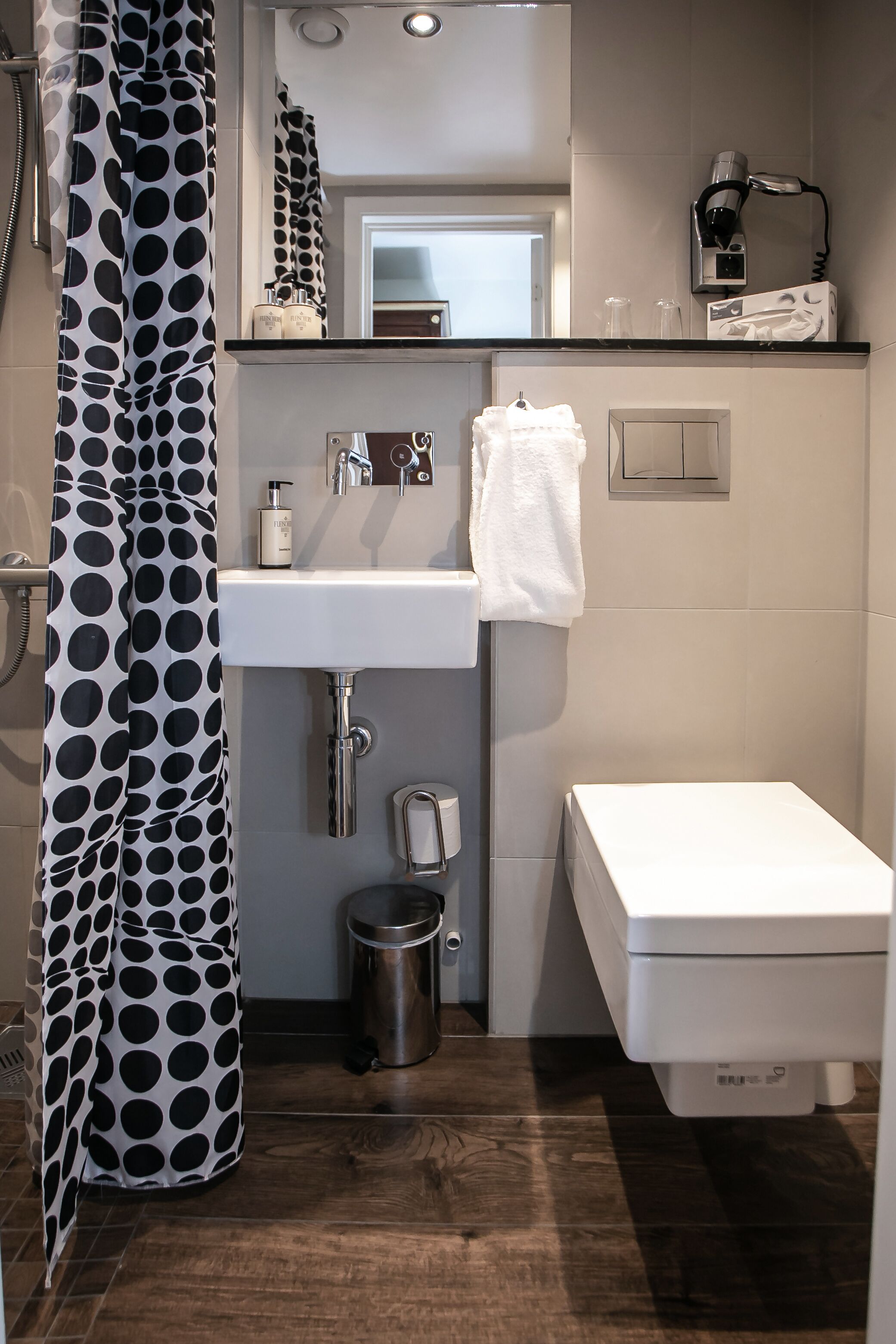 A modern bathroom with a white sink, a toilet, a shower curtain with a black and white polka dot pattern, a mirror, and a hairdryer on a shelf.
