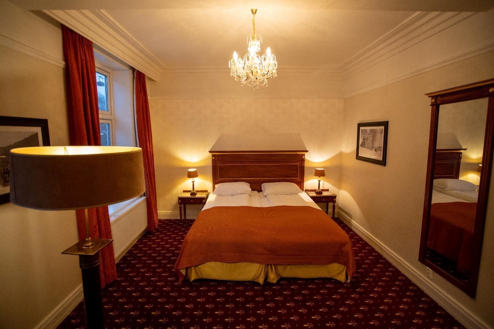 Ein Hotelzimmer mit einem großen Bett, zwei Nachttischen mit Lampen, einer Kristallleuchtre, einem Fenster mit roten Vorhängen und einem Spiegel an der Wand.