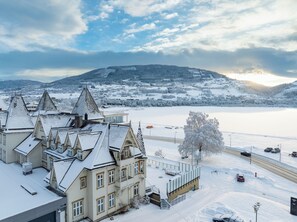 A snow-covered building with multiple roofs and a view of a snowy landscape and a lake. - Fleischer's Hotel (Voss)