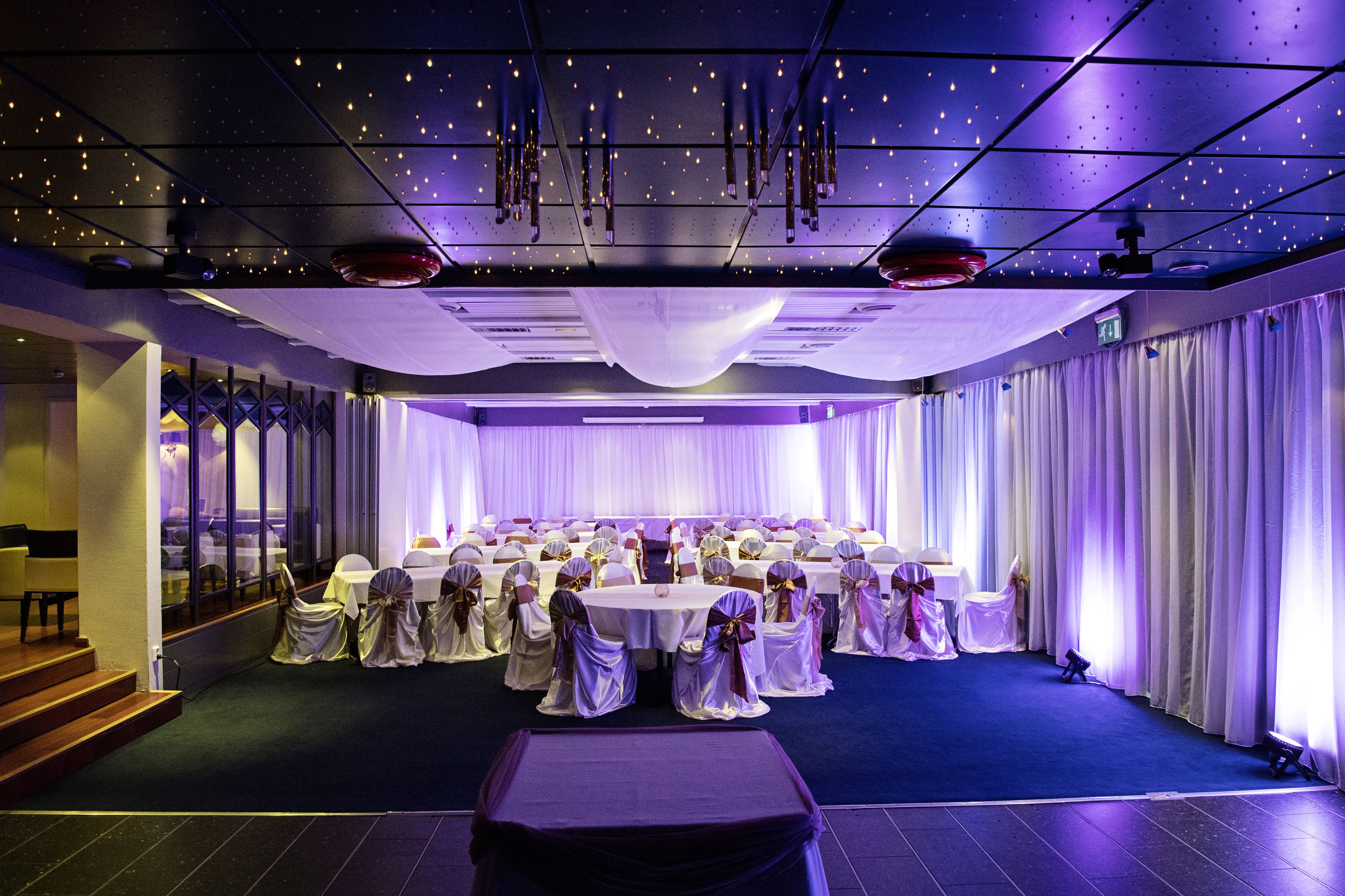 A banquet hall with round tables, chairs, and draped curtains.