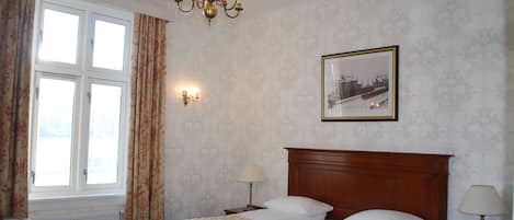Habitación de hotel con una cama grande, cabecero de madera, candelabro, un cuadro en la pared y una ventana con cortinas.