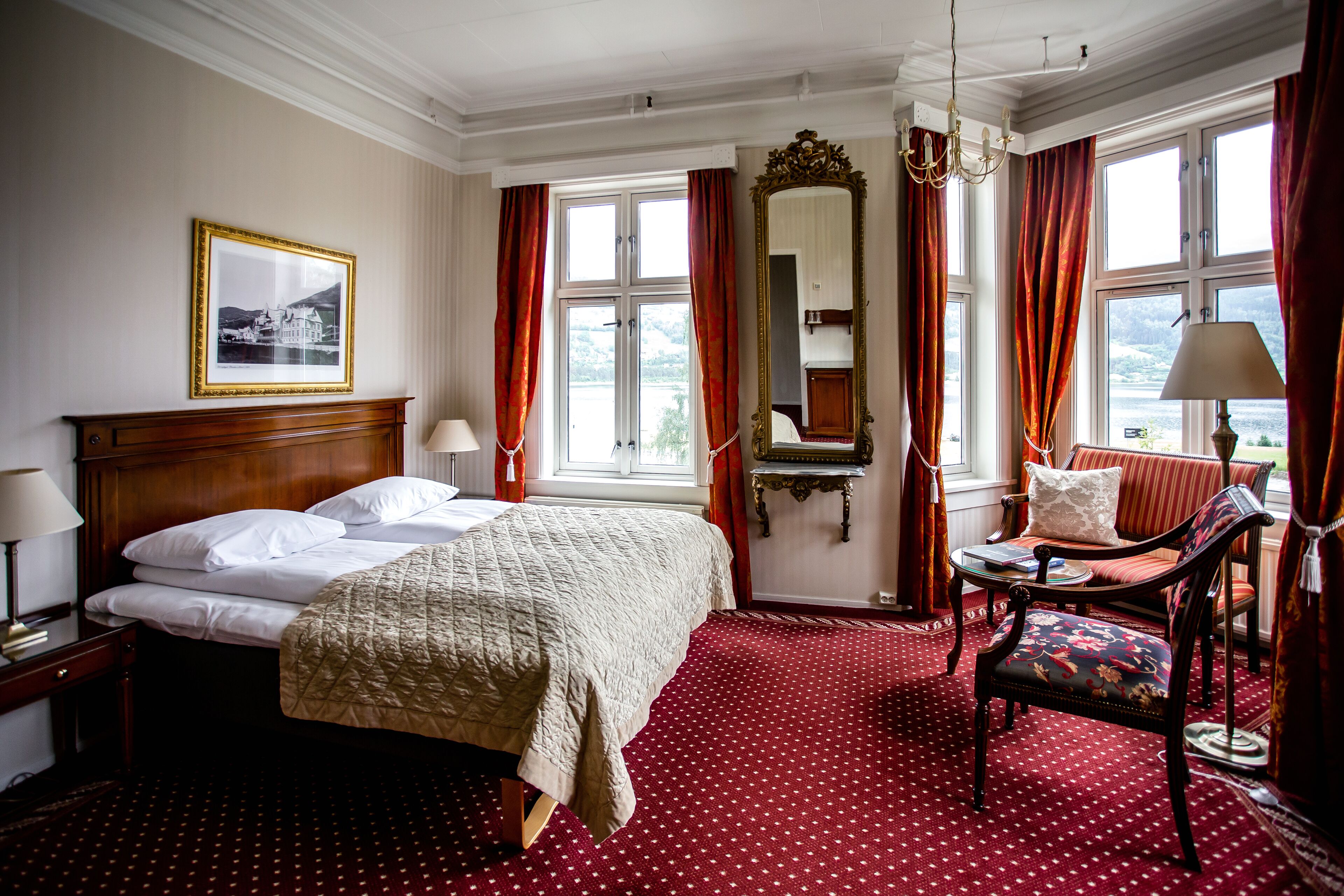 A hotel room with a large bed, a chair, a table, a mirror, and a window with curtains.