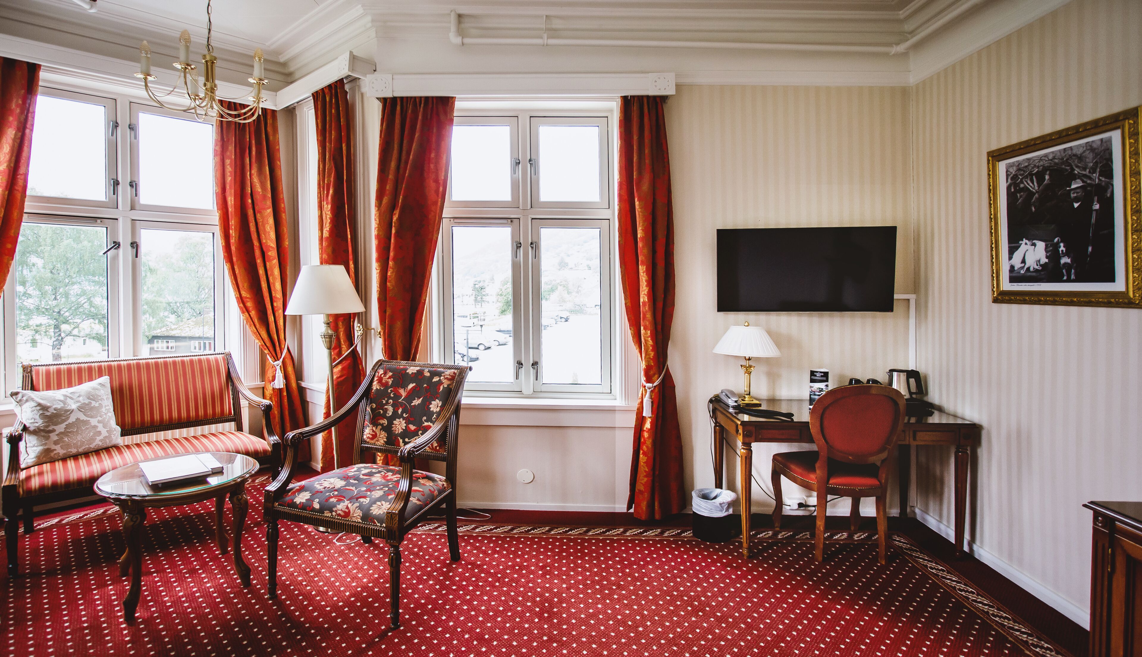 A room with a sofa, armchair, and a desk with a lamp. There is a television on the wall and a window with curtains.