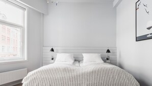 Un dormitorio minimalista con una cama grande, dos mesitas de noche, una ventana con vistas a edificios y un cuadro enmarcado en la pared.
