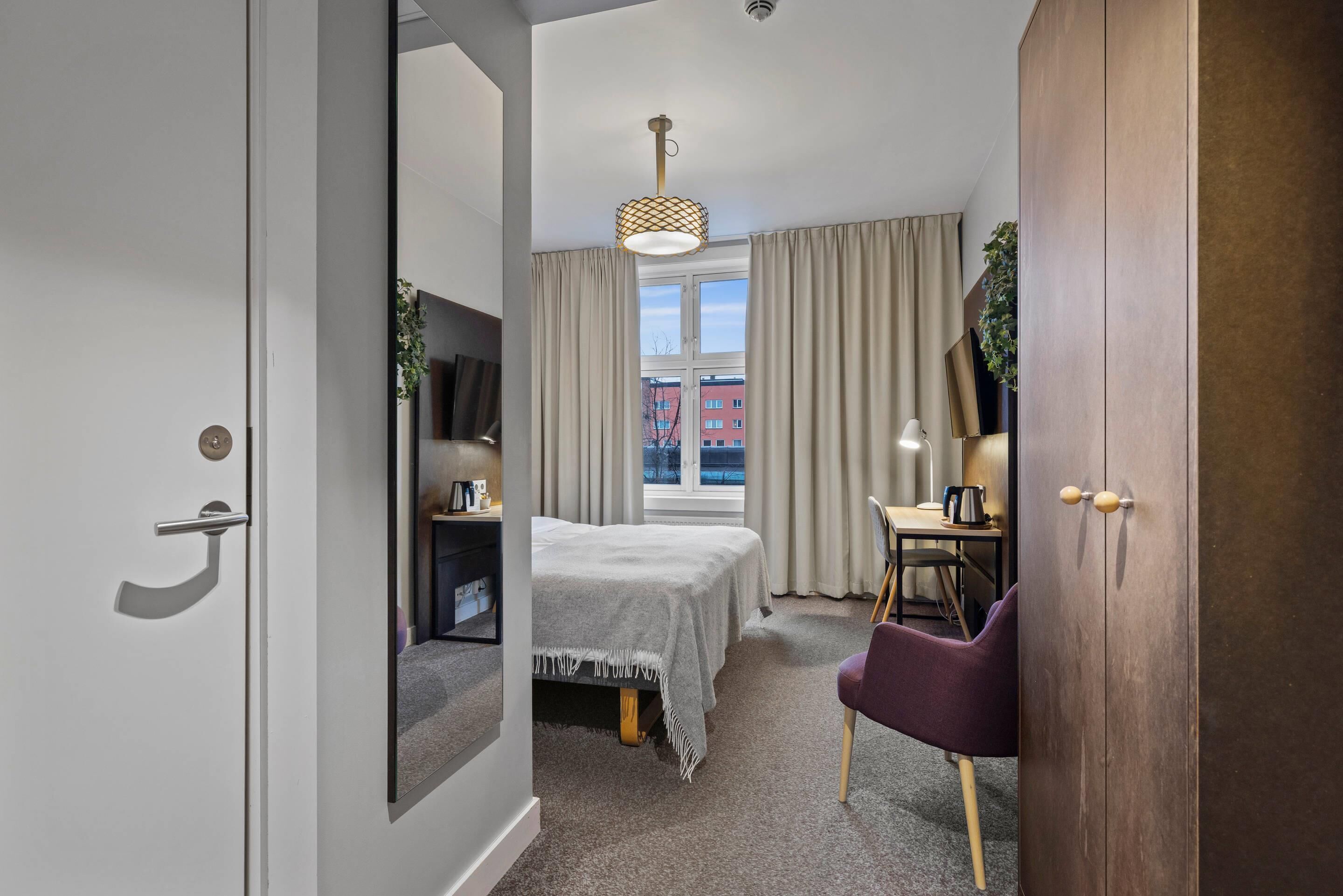 Habitación de hotel con cama, escritorio, silla, televisor y ventana con cortinas.