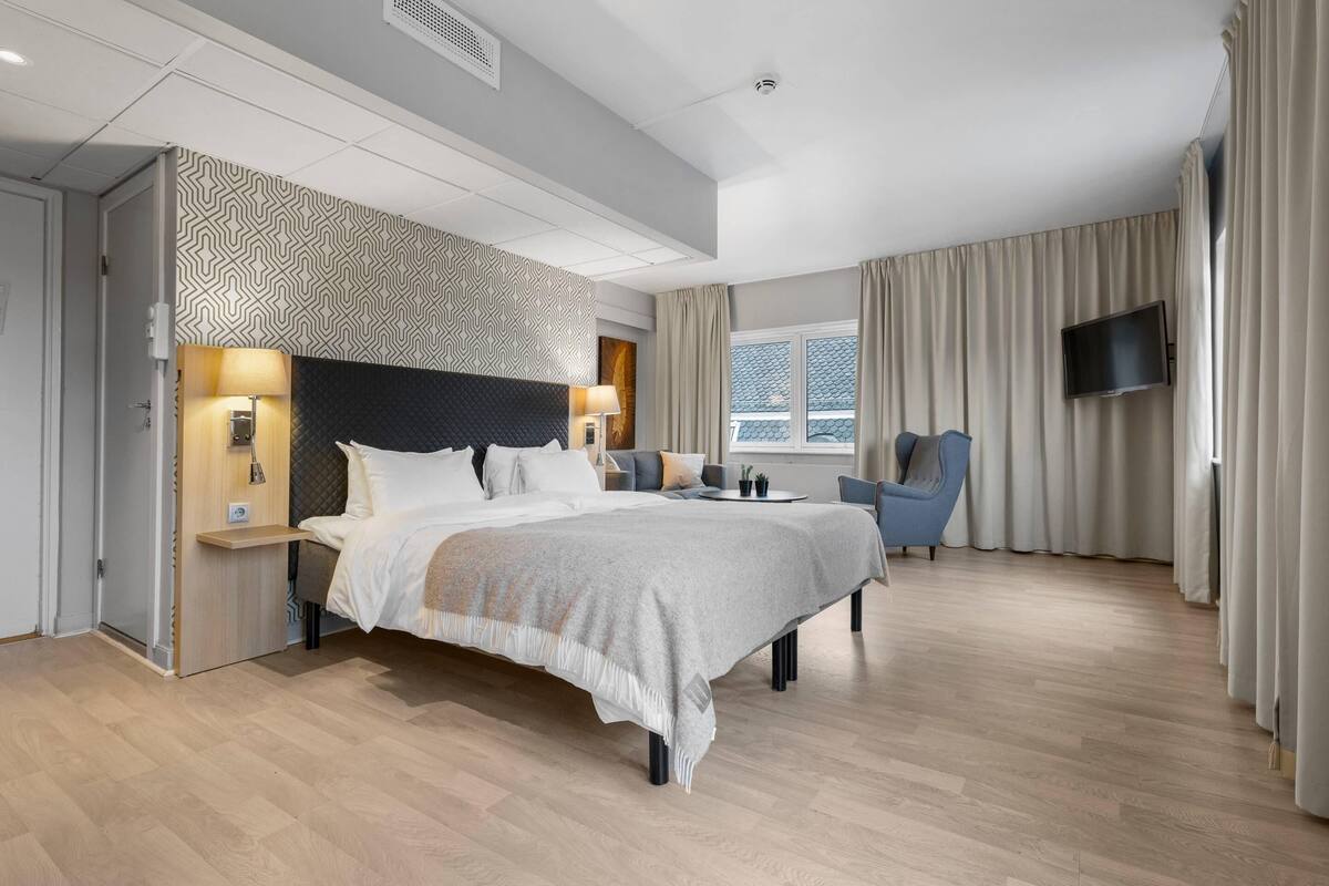 Una habitación de hotel moderna con una cama grande, dos mesitas de noche, un televisor y una ventana con cortinas.