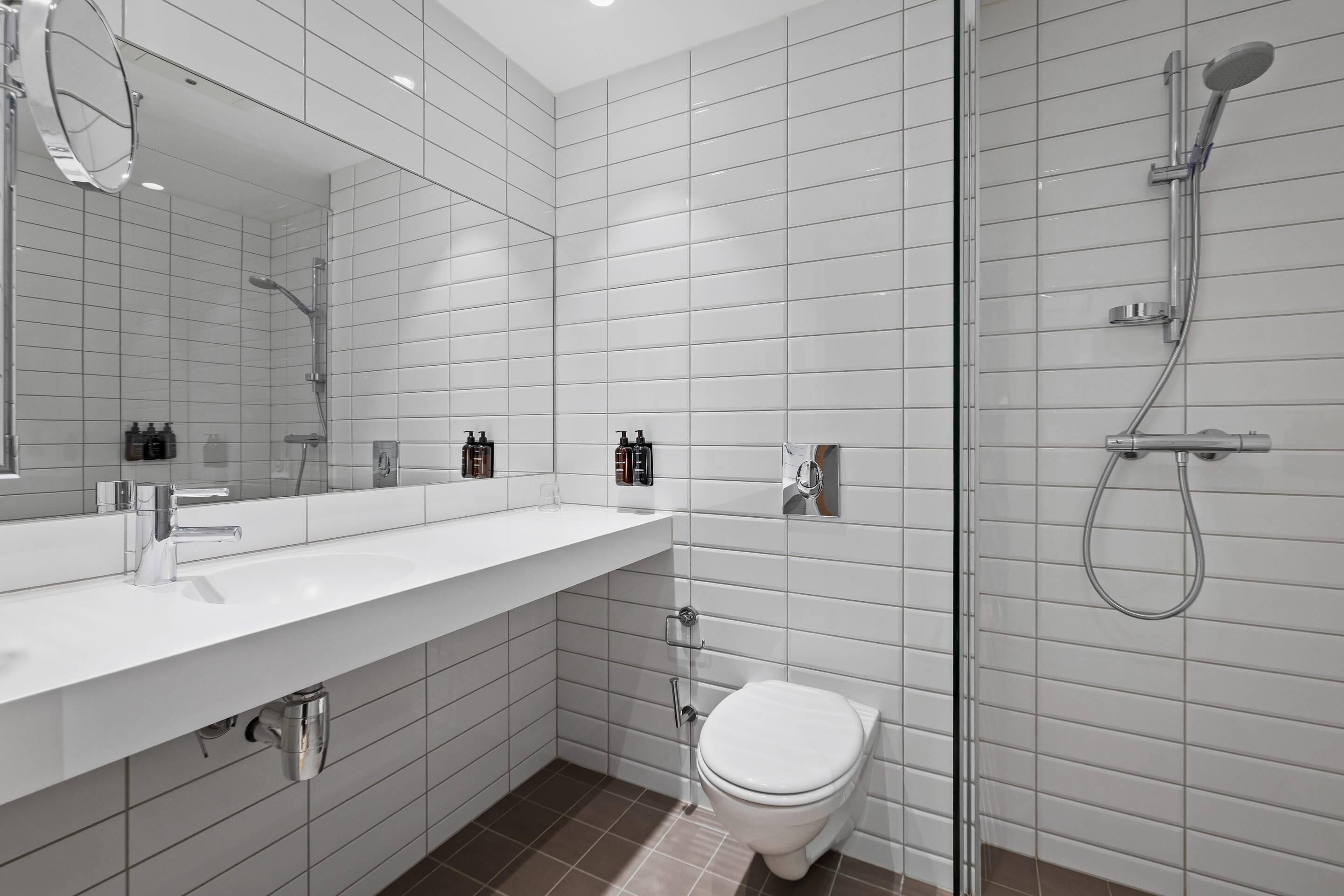 Un baño moderno con pared de azulejos blancos, un espejo grande, un lavabo blanco, un inodoro y una zona de ducha.