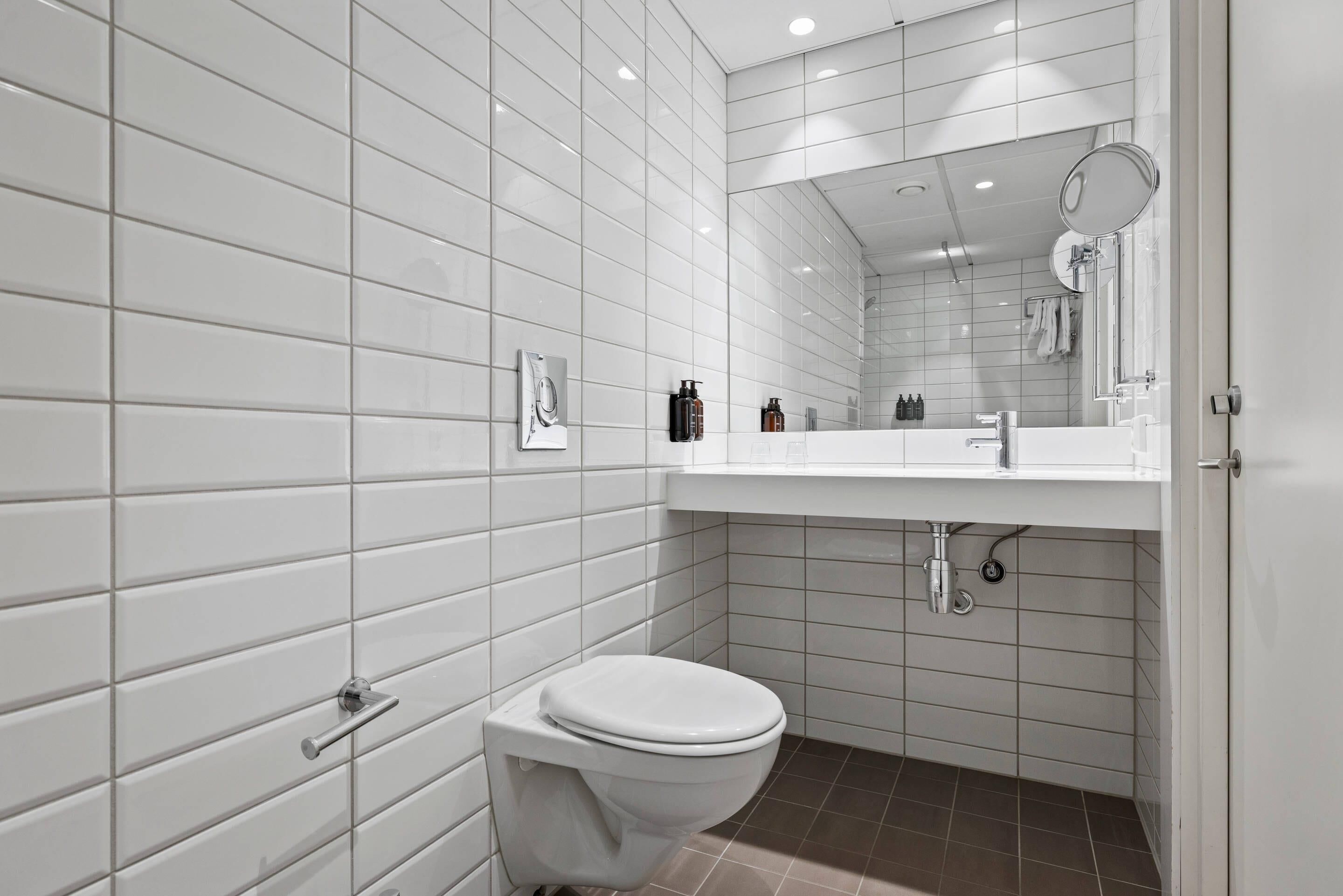 A modern bathroom with white tiled walls, a white toilet, a sink with a mirror, and a shower area.