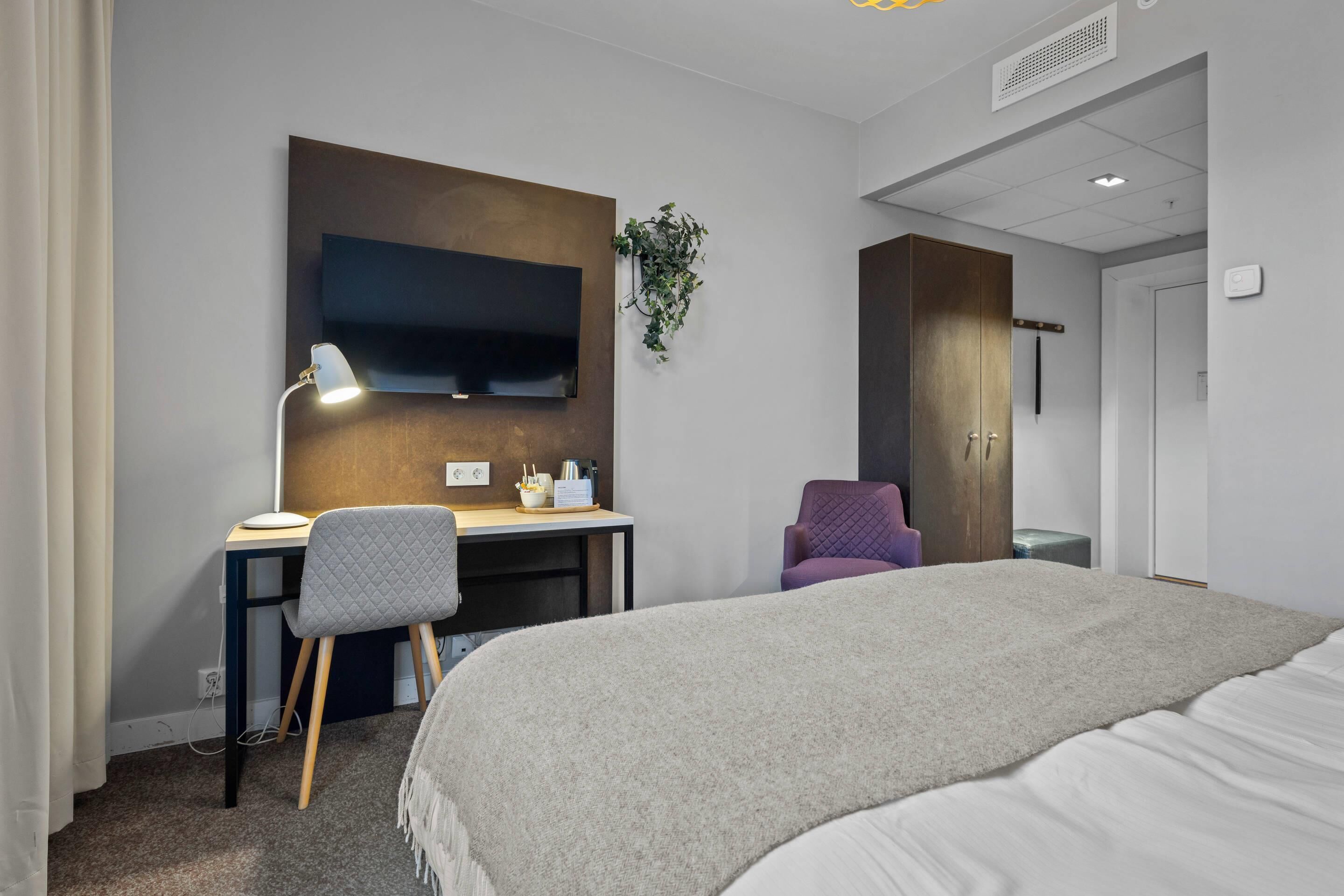 Ein modernes Hotelzimmer mit einem Bett, einem Schreibtisch mit Stuhl, einem an der Wand montierten Fernseher und einer kleinen Pflanze.