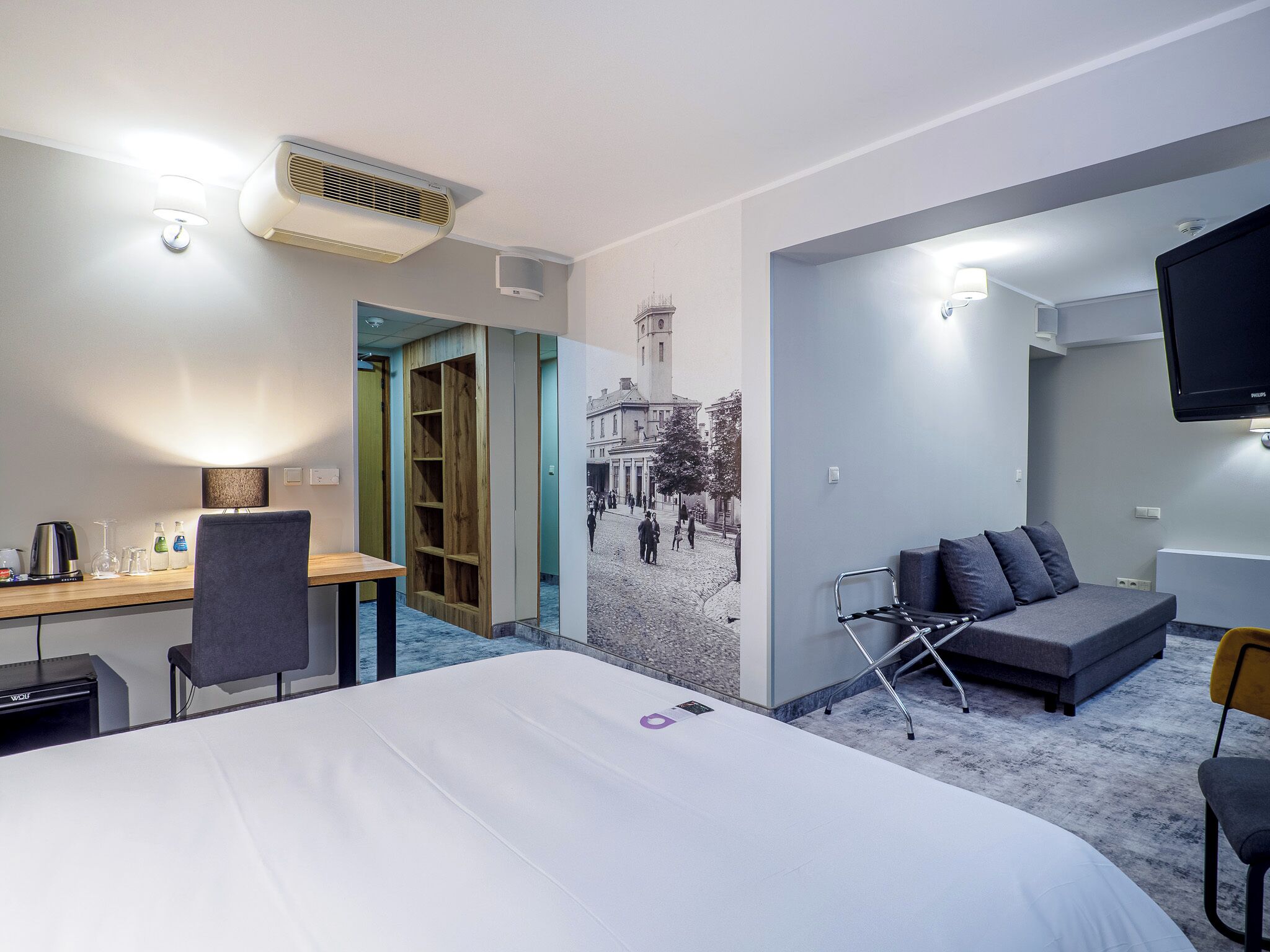 Habitación de hotel con cama, escritorio con lámpara, televisor, sofá y pared con un mural de paisaje urbano.