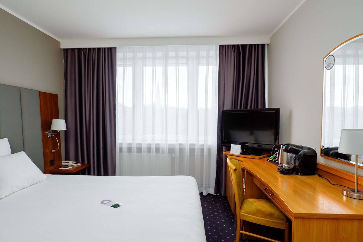 A hotel room with a bed, a desk with a television, a chair, a desk lamp, and a window with curtains.