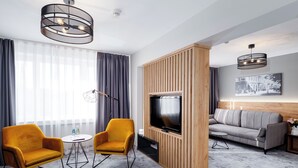 Una habitación de hotel moderna con televisión, dos sillones amarillos, un sofá gris y un separador de madera.