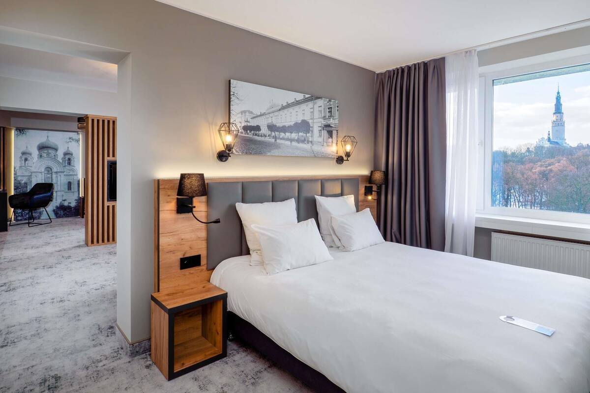 A hotel room with a large bed, a wooden nightstand, a wall-mounted lamp, and a view of a cityscape through a window.