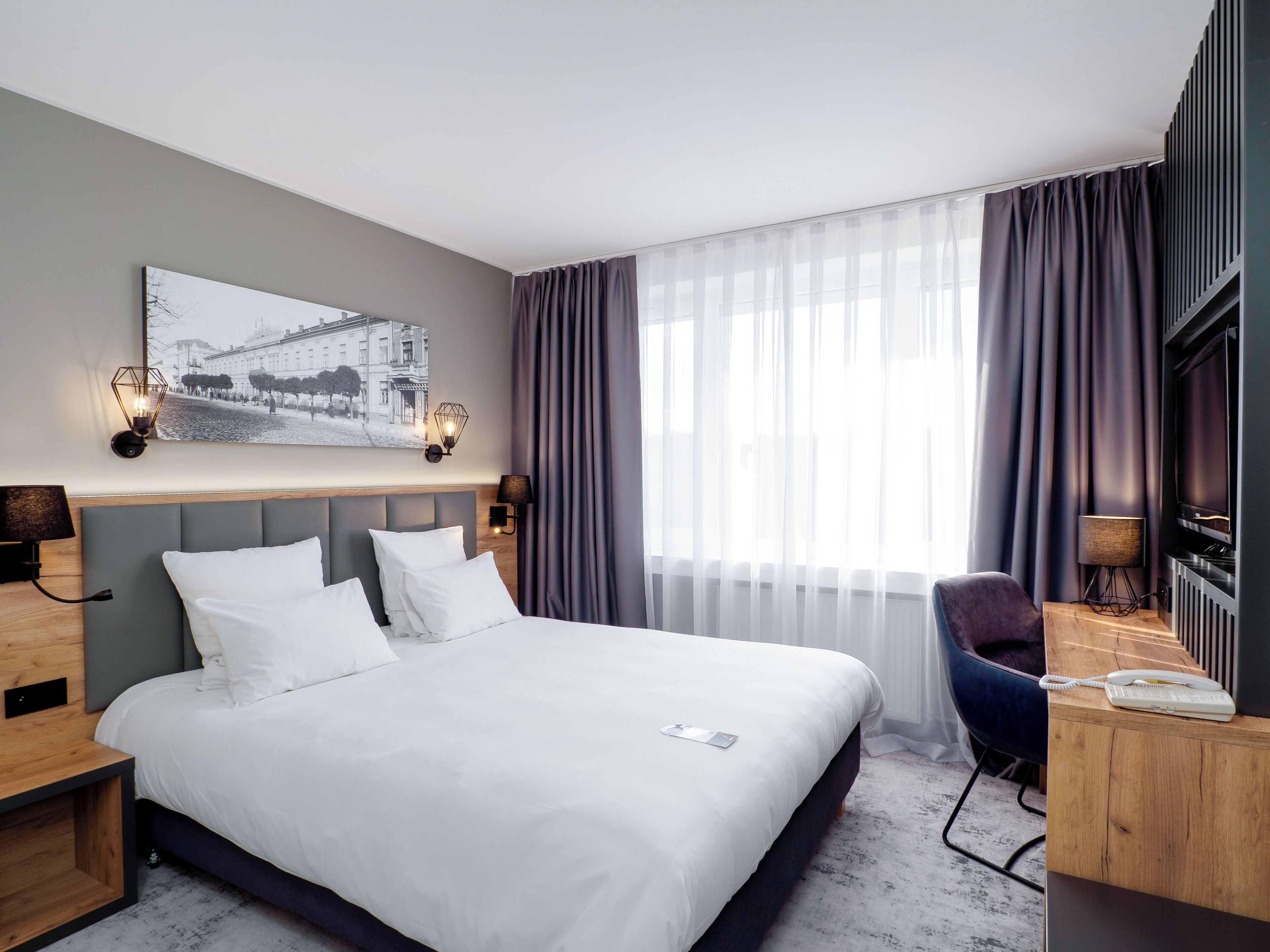 A modern hotel room with a large bed, a desk with a telephone, a chair, and a window with curtains.