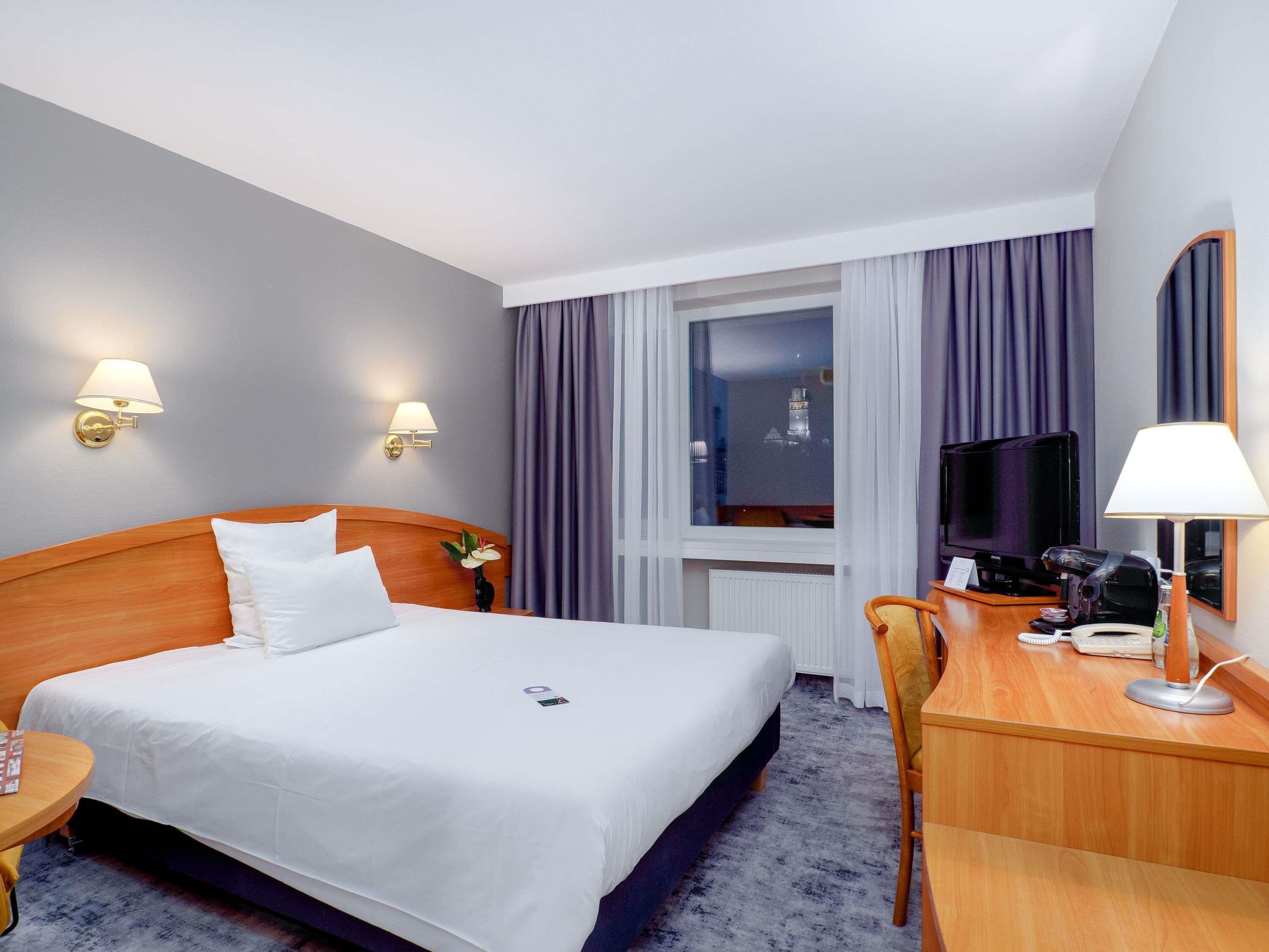 Une chambre d’hôtel avec un lit, un bureau avec un ordinateur, une télévision et une fenêtre avec des rideaux.