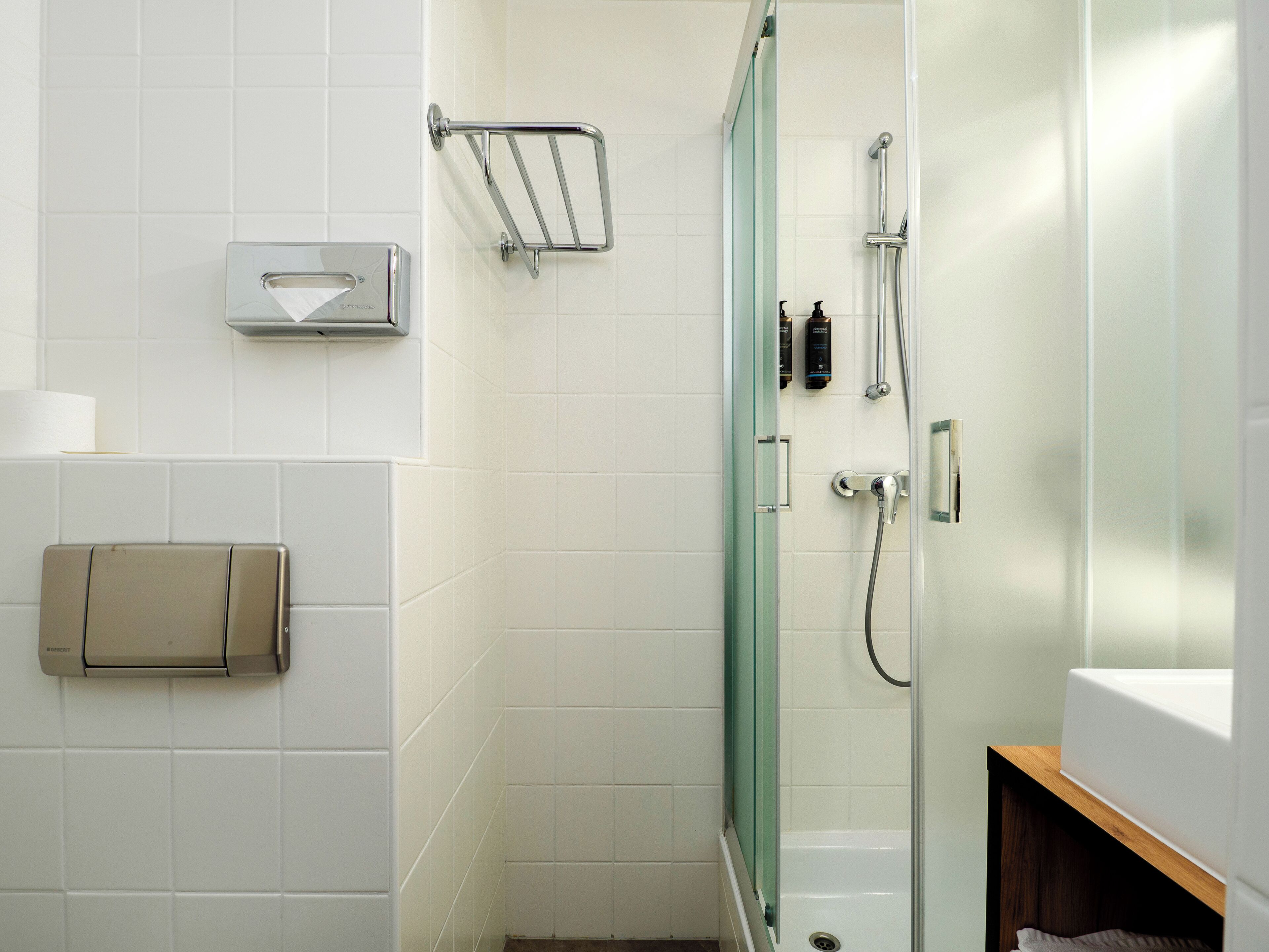 Et moderne badeværelse med hvide fliser, en glasbrusekabine, toiletpapirholder og en vask med træbordplade.