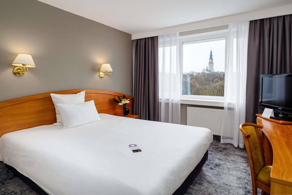 A hotel room with a bed, bedside lamps, a television, a desk, and a window with a view of a building.