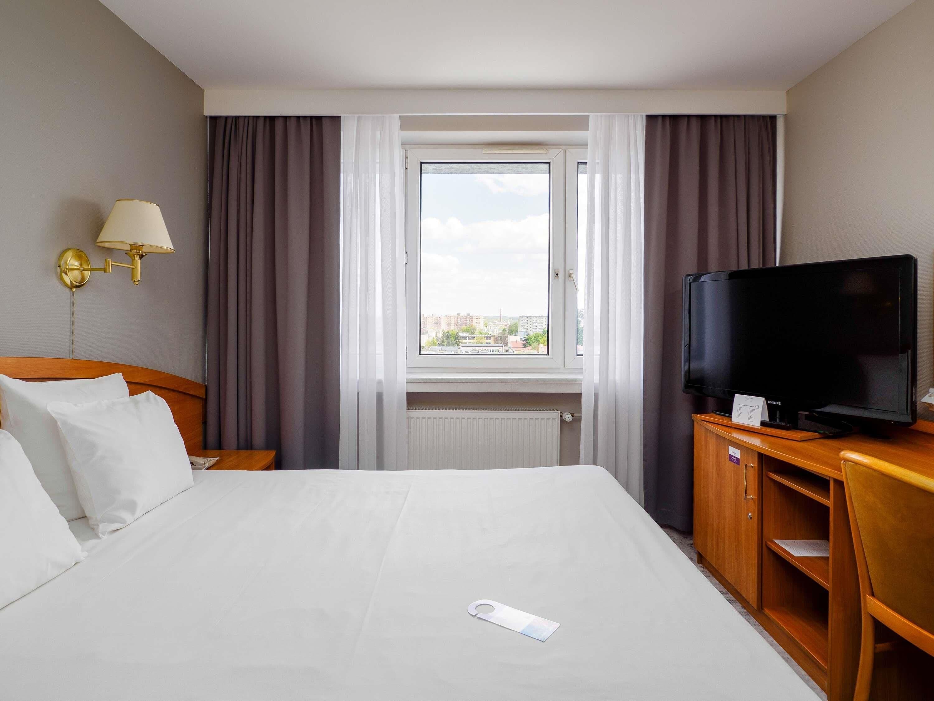 Et hotelværelse med seng, fjernsyn, lampe og et vindue med gardiner.
