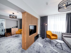 Ein modernes Wohnzimmer mit Fernseher, einer Wand in Holzoptik, zwei gelben Sessel, einem grauen Sofa und einem großen Fenster mit Vorhängen.