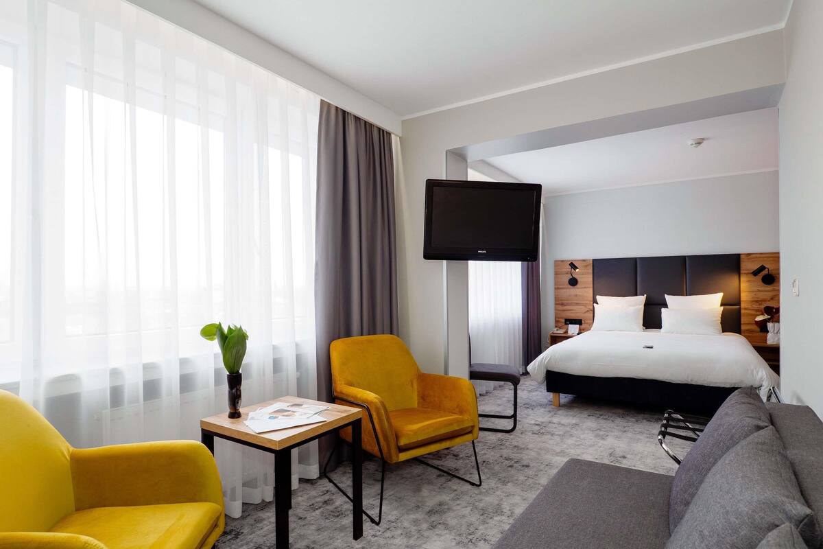 A hotel room with a bed, a TV, a sofa, a chair, a small table, and a plant.
