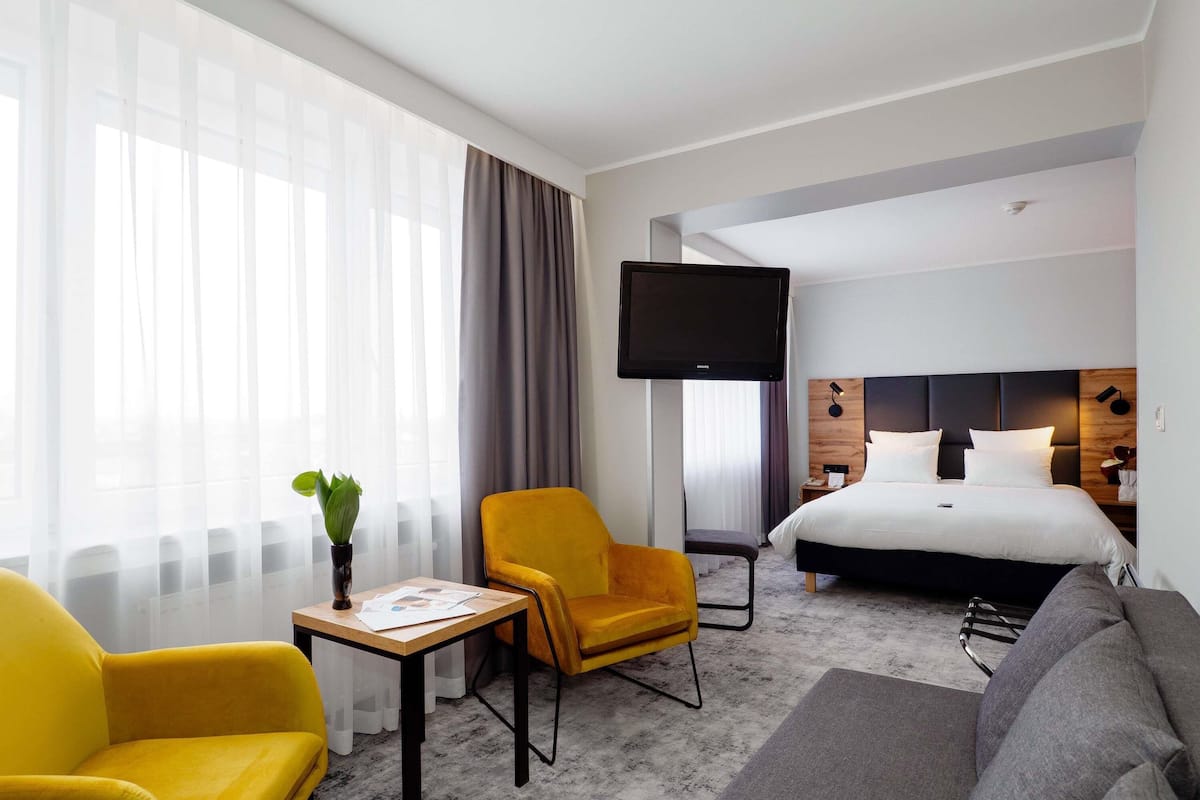 A hotel room with a bed, a TV, a sofa, a chair, a small table, and a plant.