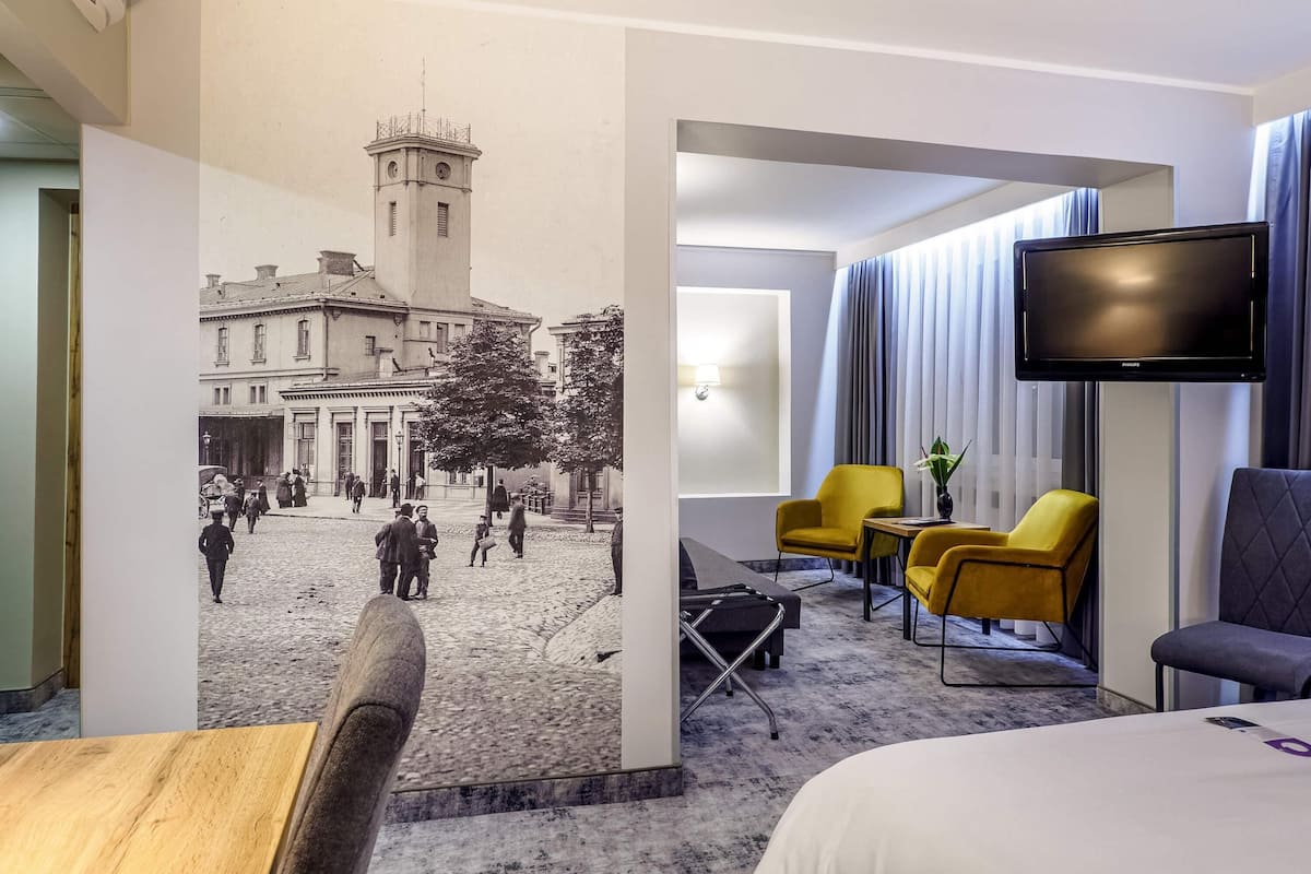 A hotel room with a bed, a flat-screen TV, a desk, a chair, and a wall with a historical photo.