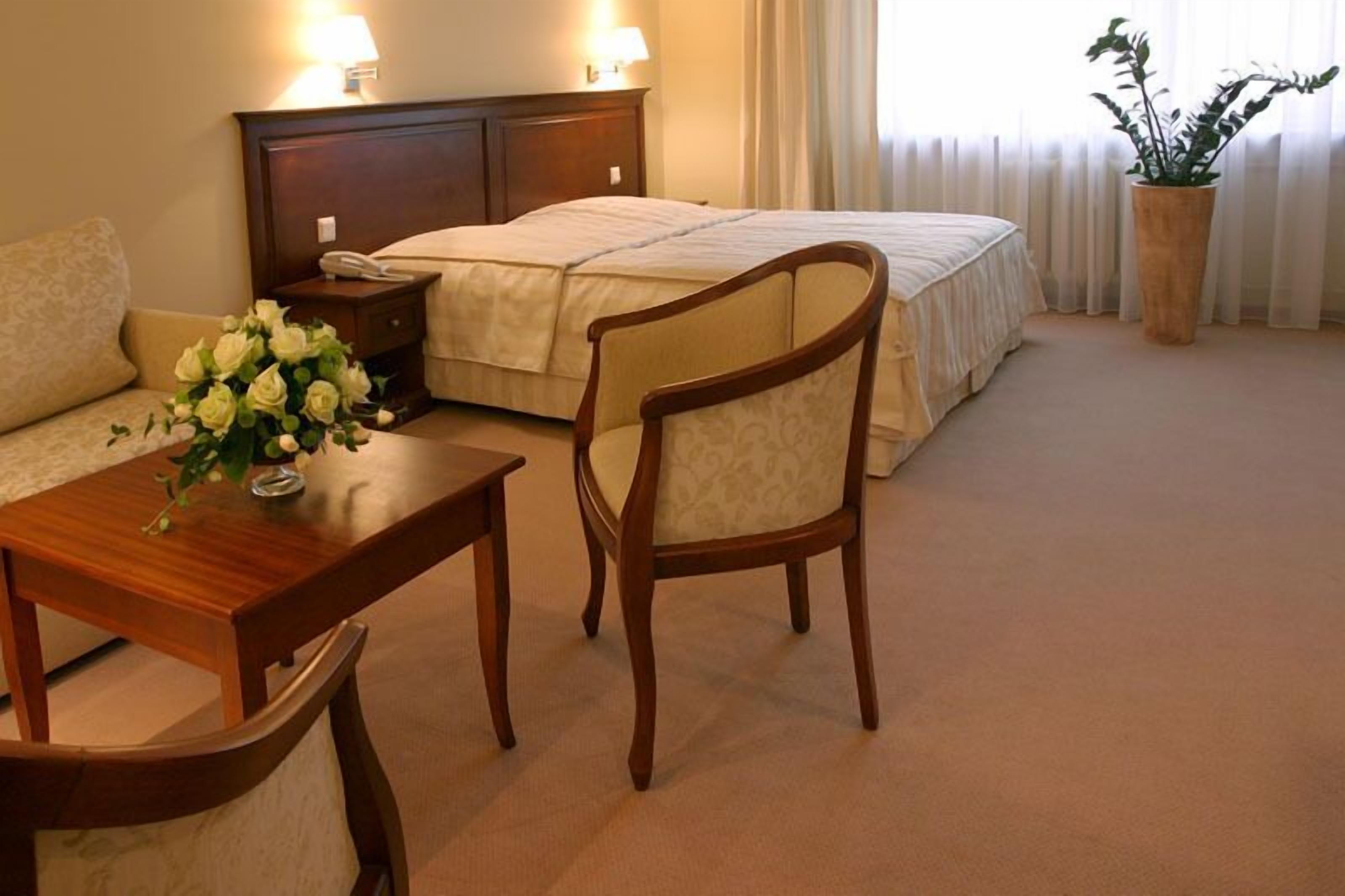 A hotel room with a bed, a wooden nightstand, a bedside table with a vase of flowers, a wooden chair, and a potted plant.