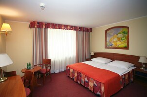 Una habitación de hotel con una cama, un escritorio con una lámpara, una silla, una mesa con dos botellas y un cuadro en la pared.