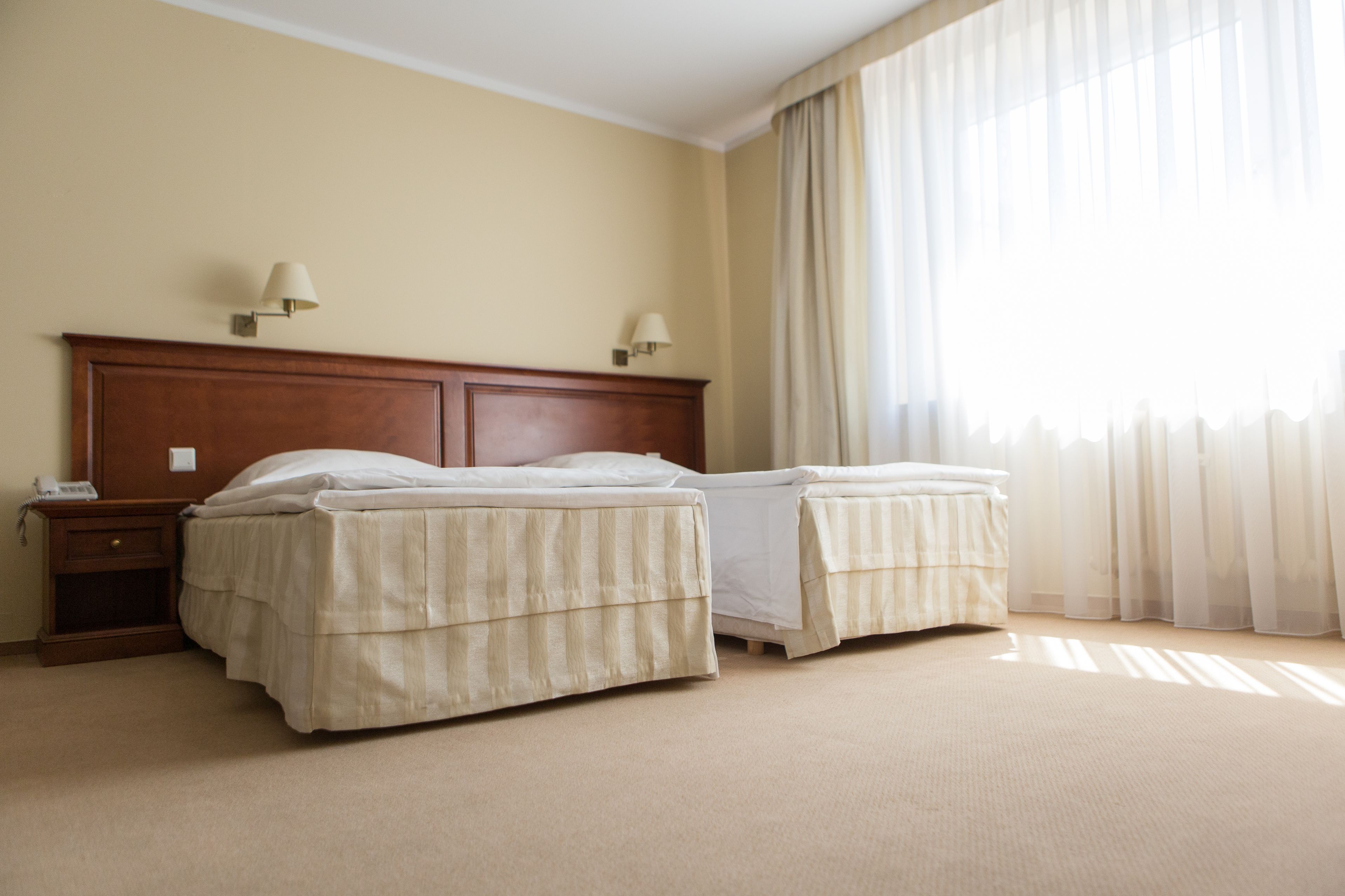 A hotel room with two beds, a wooden headboard, bedside lamps, a nightstand, and a large window with sheer curtains.