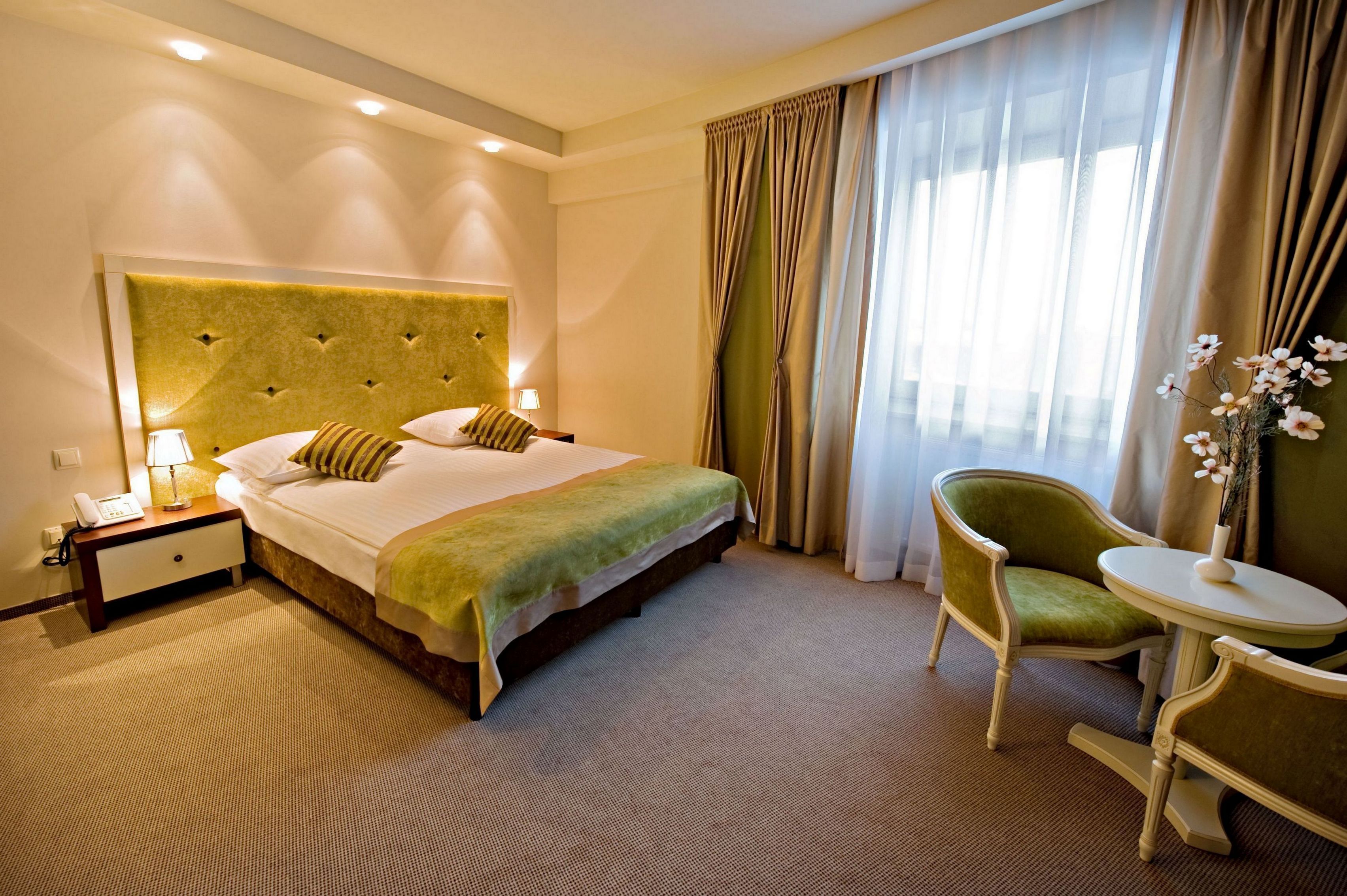 Una habitación de hotel con una cama grande, una mesita de noche con lámpara, un sillón verde, una pequeña mesa redonda y un florero con flores.