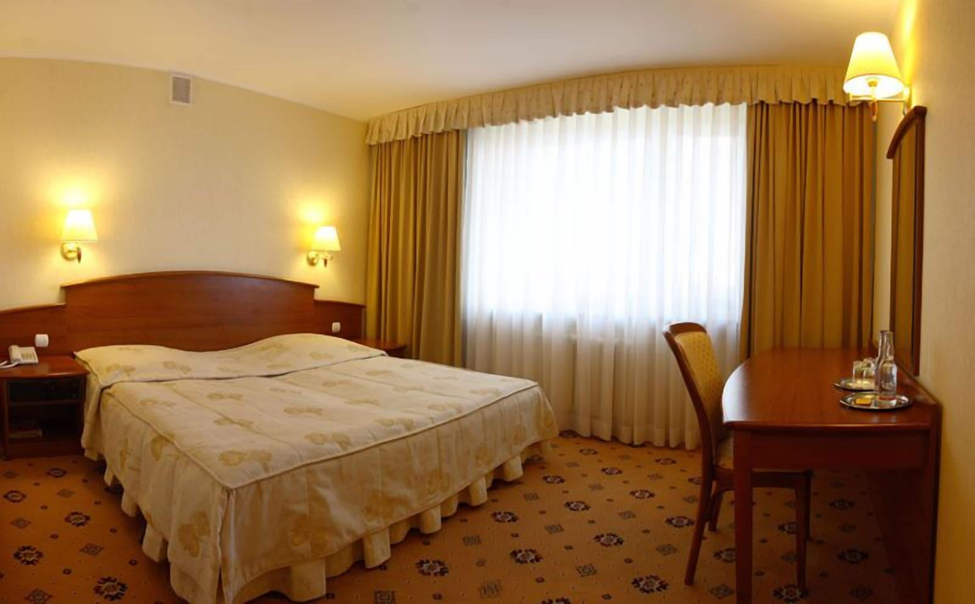 A hotel room with a double bed, a bedside table with a lamp, a chair, and a large window with curtains.