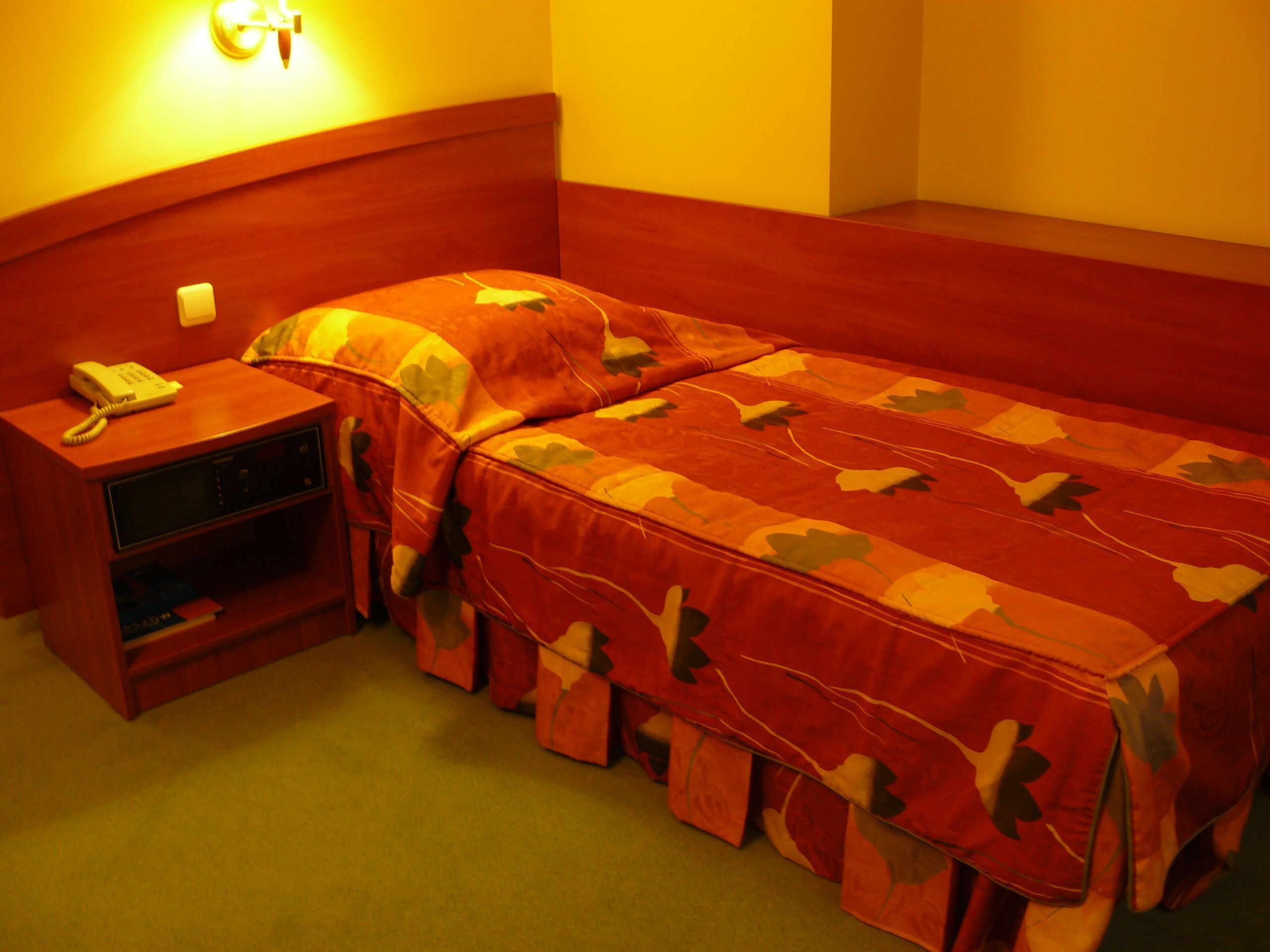 A single bed with a red leaf-patterned bedspread, a bedside table with a telephone, and a wall-mounted light fixture.
