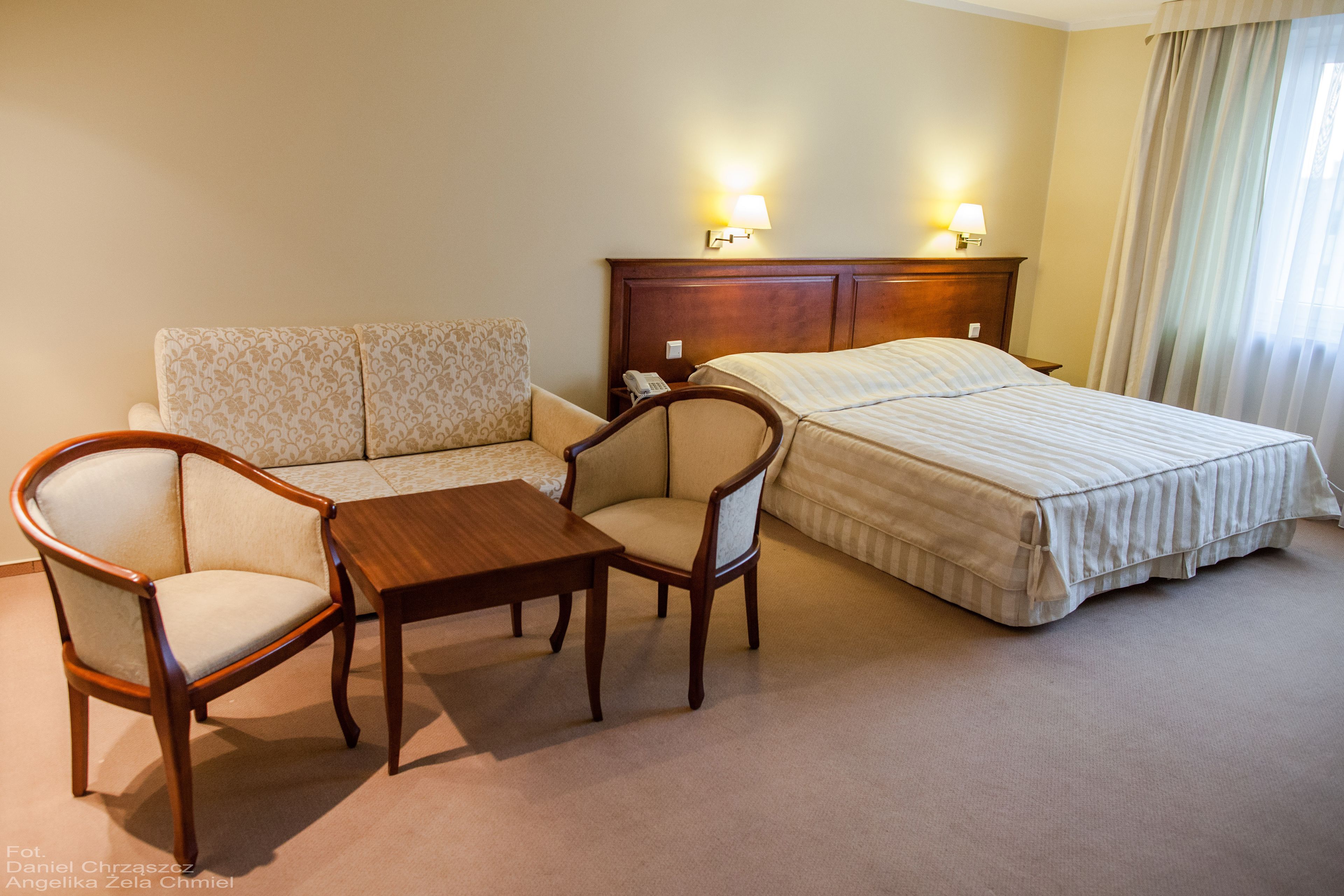 A hotel room with a bed, two armchairs, a small table, and a sofa. There are two wall-mounted lamps on the bed's headboard.