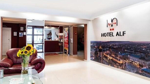 A hotel lobby with a reception desk, a glass table with a vase of sunflowers, a leather sofa, and a large wall mural of a cityscape. - Hotel Alf (Krakow)