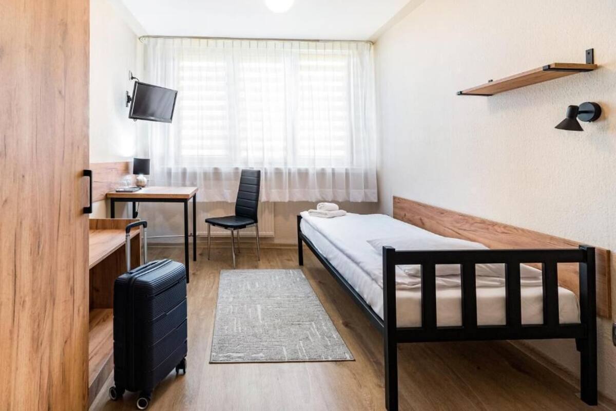Une chambre d’hôtel de taille réduite, comprenant un lit simple, un bureau avec une chaise, un téléviseur fixé au mur et une valise posée sur le sol.