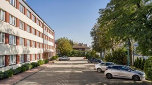 A parking area with cars, a building with red and white facade, and trees. - Hotel Alf (Krakow)