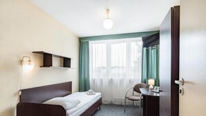 A small, neatly arranged hotel room with a single bed, a chair, a desk with a lamp, and a window with sheer curtains. - Hotel Alf (Krakow)