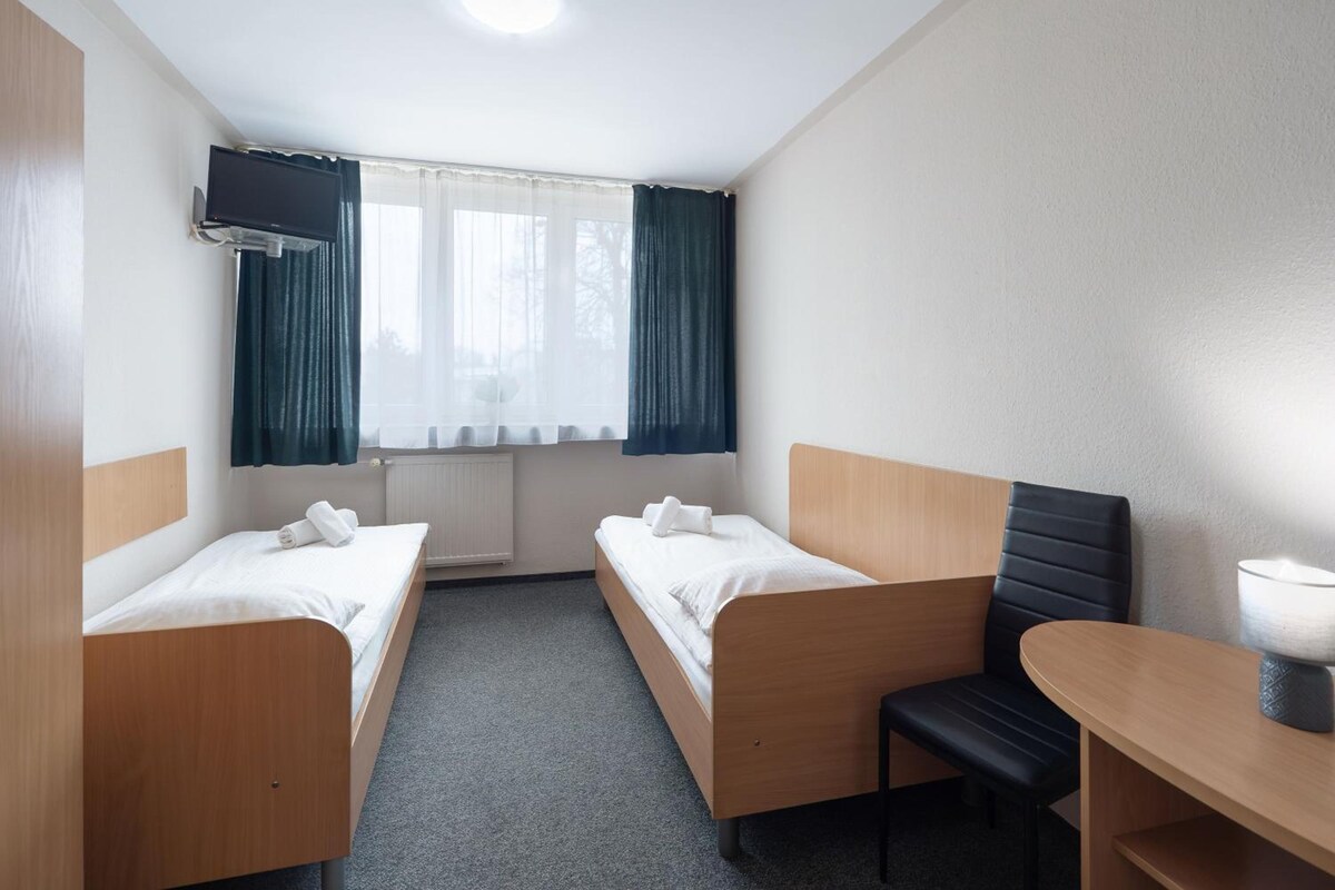 Une chambre d’hôtel avec deux lits simples, une petite table, une chaise, un téléviseur fixé au mur et une fenêtre avec des rideaux.