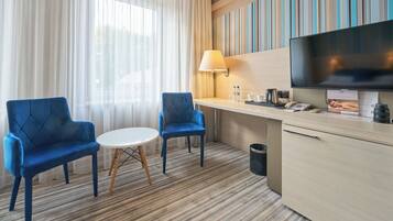 Ein Hotelzimmer mit zwei blauen Sesseln, einem kleinen runden Tisch, einem Fernseher, einer Lampe, einer Minibar und einem großen Fenster mit transparenten Vorhängen.