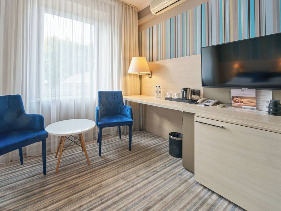Ein Hotelzimmer mit zwei blauen Sesseln, einem kleinen runden Tisch, einem Fernseher, einer Lampe, einer Minibar und einem großen Fenster mit transparenten Vorhängen.