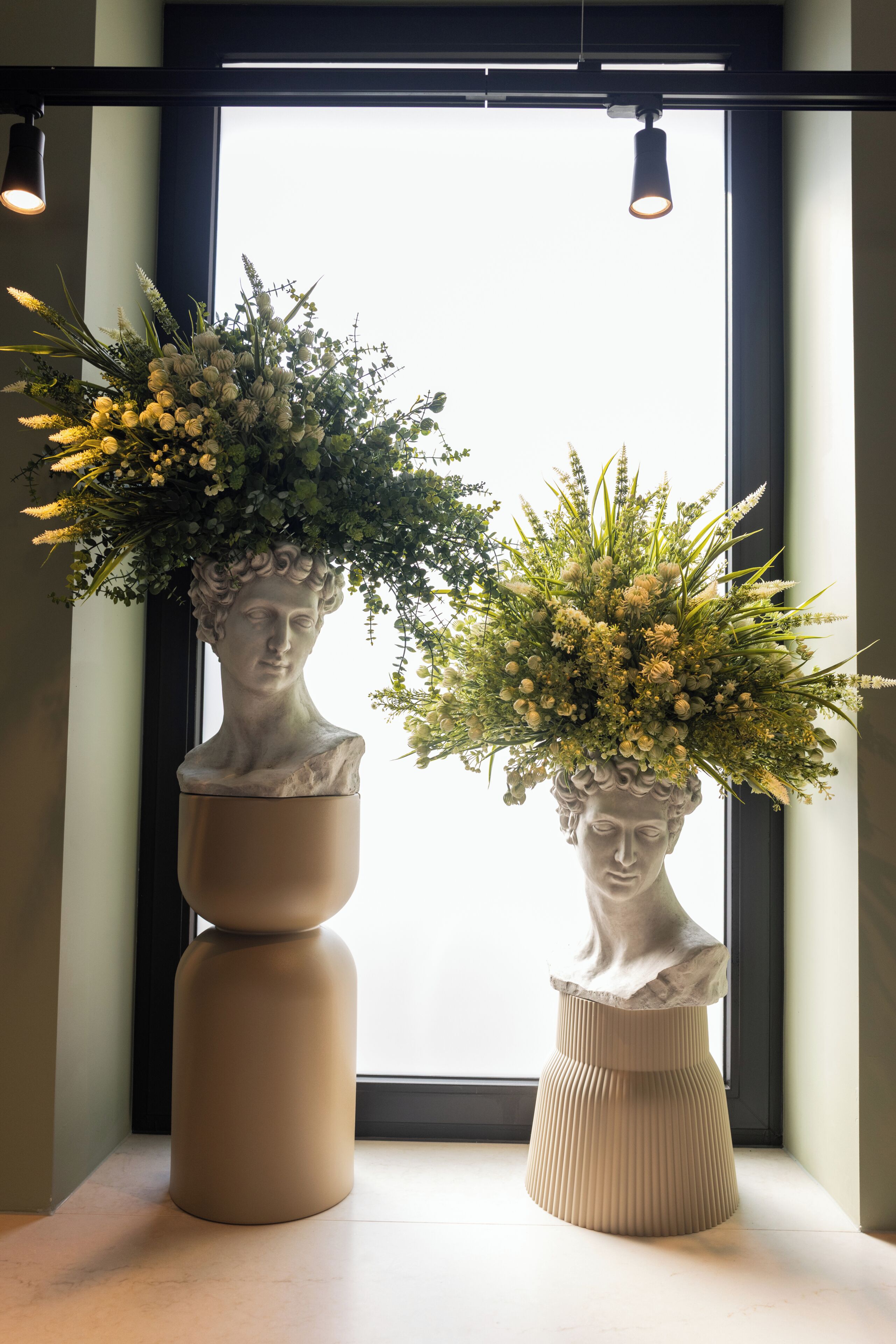Dos esculturas de bustos con arreglos florales frente a una ventana.