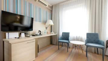 Habitación de hotel con televisión de pantalla plana, escritorio con lámpara, dos sillas, una mesita y un gran ventanal con cortinas.
