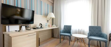 A hotel room with a flat-screen TV, a desk with a lamp, two chairs, a small table, and a large window with curtains.