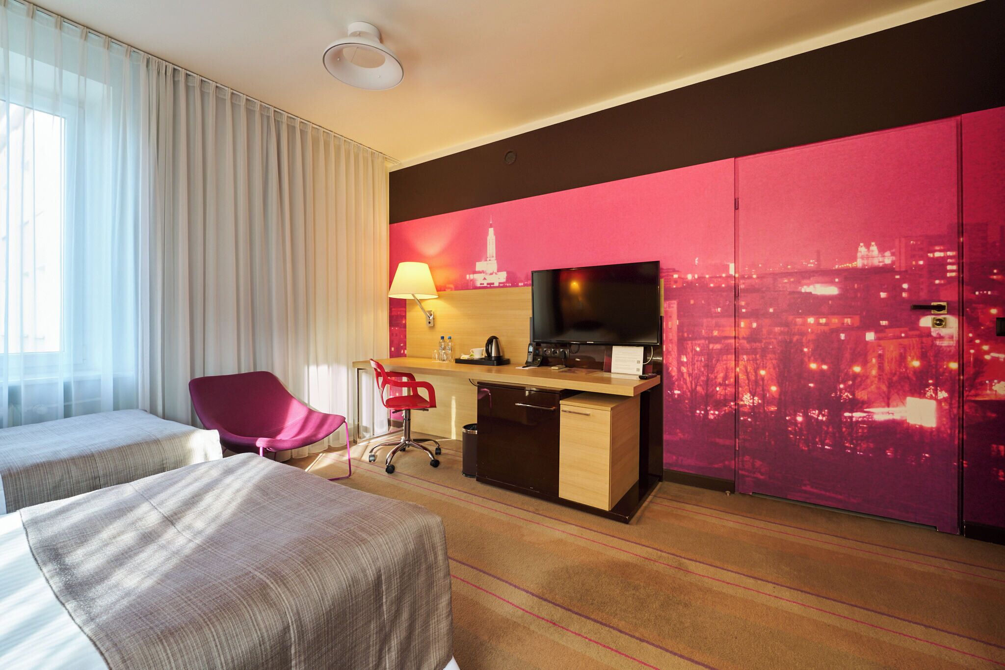 Habitación de hotel con dos camas, un escritorio con televisor, una silla, una lámpara y vista panorámica de la ciudad.