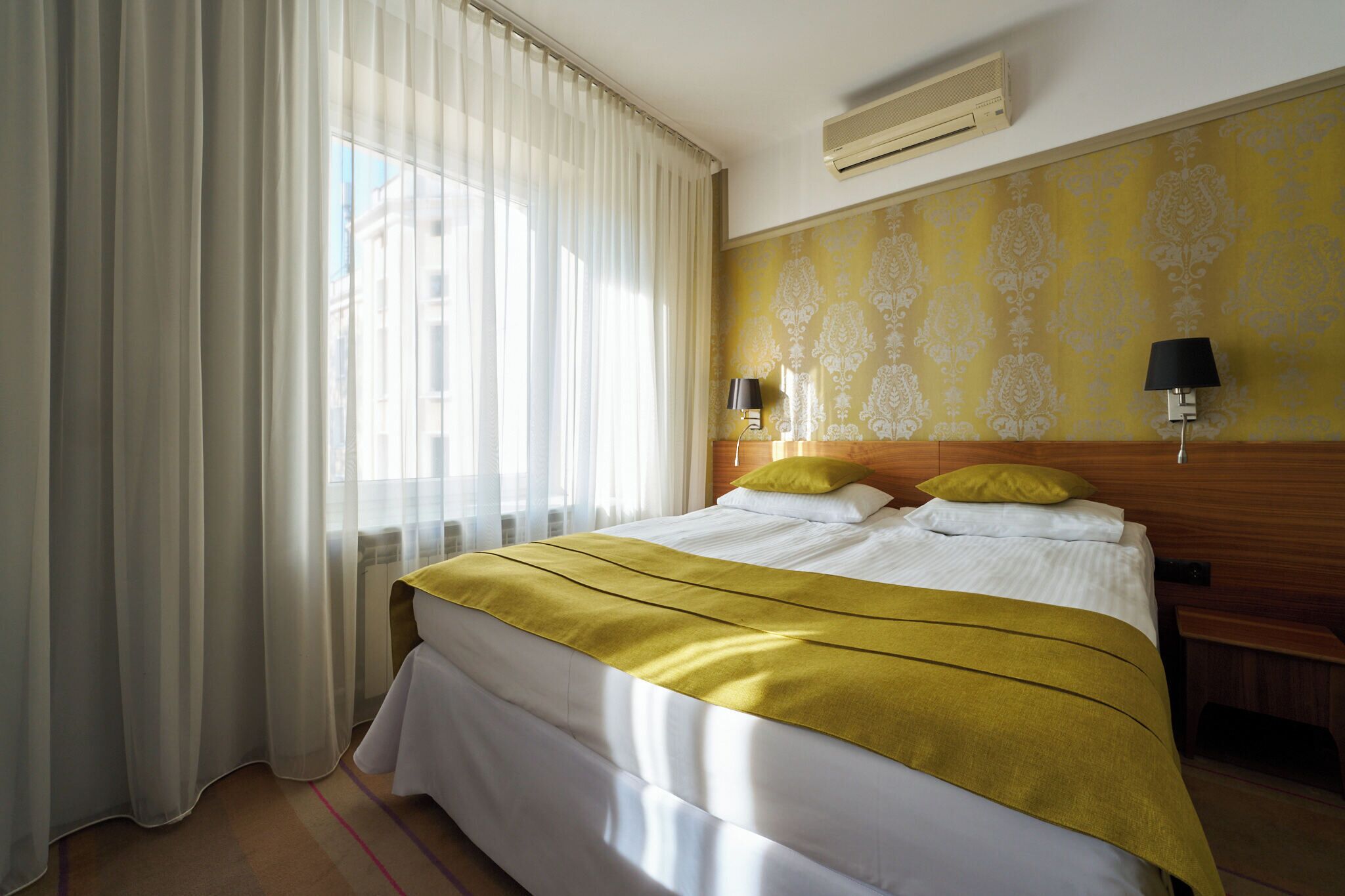 A hotel room with a bed, curtains, a wall with a pattern, a nightstand, and a lamp.