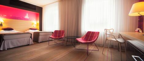 A hotel room with two beds, a small round table, two armchairs, a desk with a lamp, and a large window with curtains.
