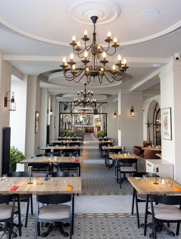 A spacious restaurant with multiple dining tables, a grand chandelier, and a view into the dining area.