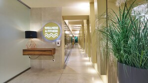 A hallway with a wellness and fitness sign, a wooden bench, a wall-mounted soap dispenser, and a large planter with green plants. - Hotel Cristal (Bialystok)
