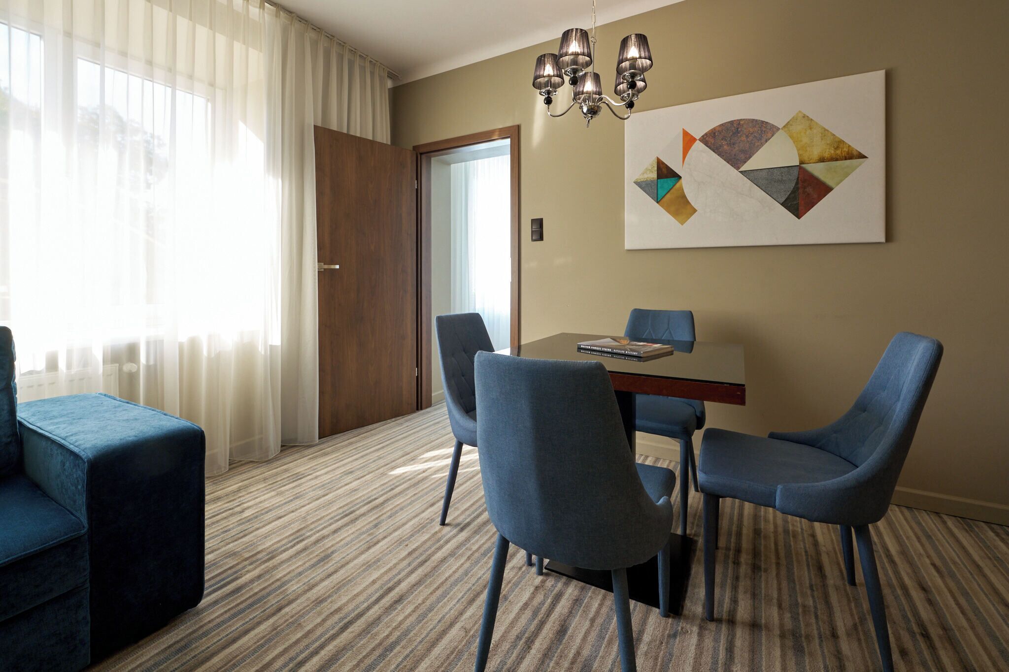 A hotel room with a dining area, a blue sofa, a chandelier, and a geometric artwork on the wall.