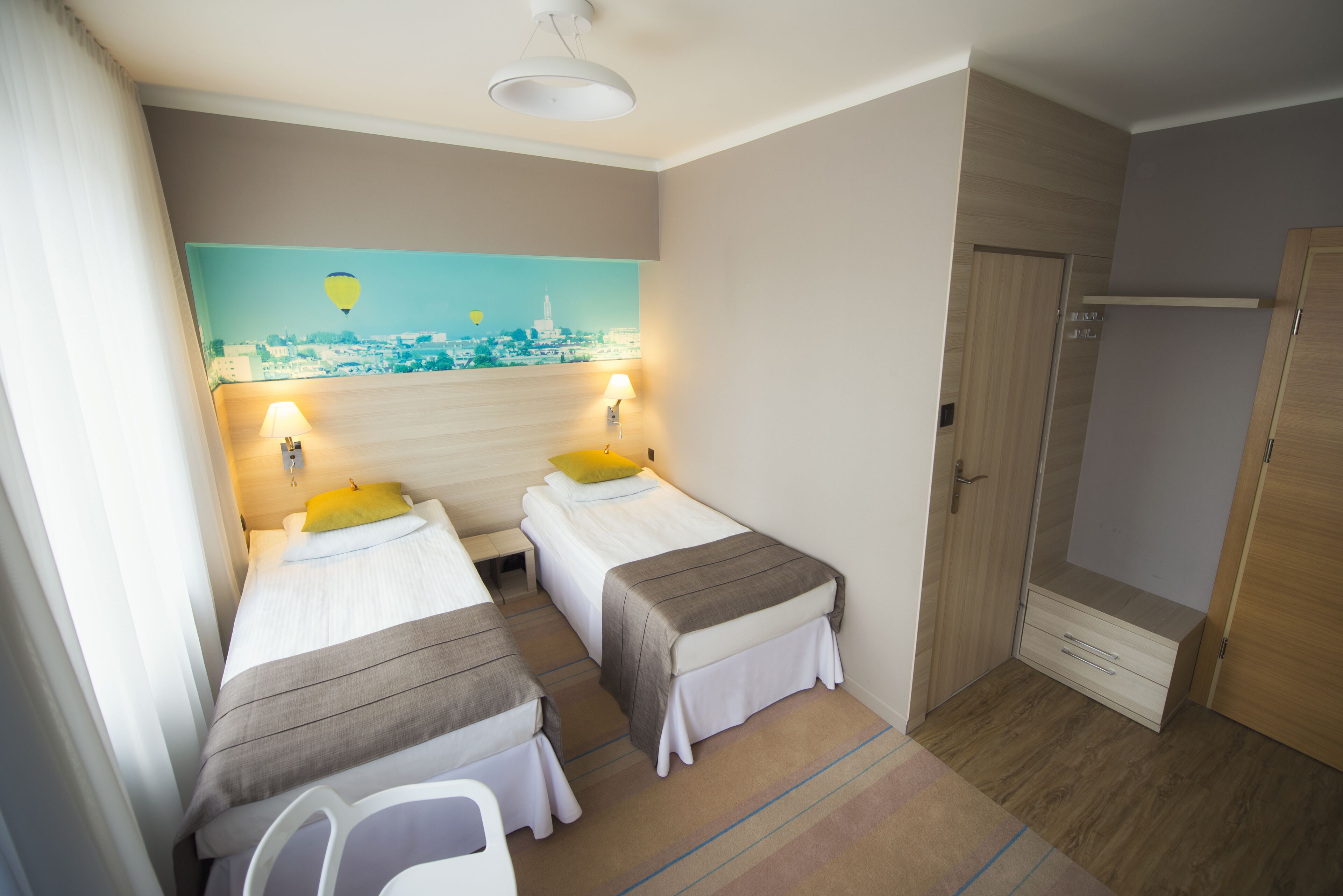 A hotel room with two single beds, a painting of a cityscape, a wooden wardrobe, and a shower area.